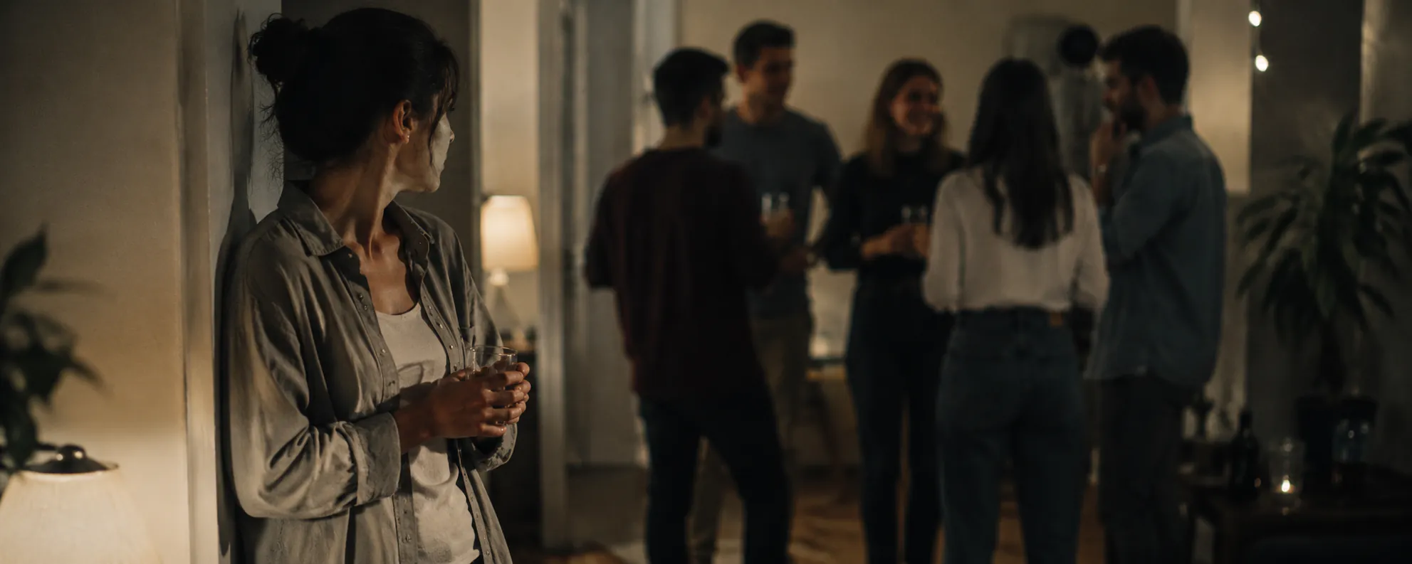 Frau lehnt zurückhaltend an der Wand abseits einer geselligen Runde bei einer Feier – Symbolbild Unsicherheit im sozialen Kontakt, Hypno-Flirt-Coaching in Augsburg